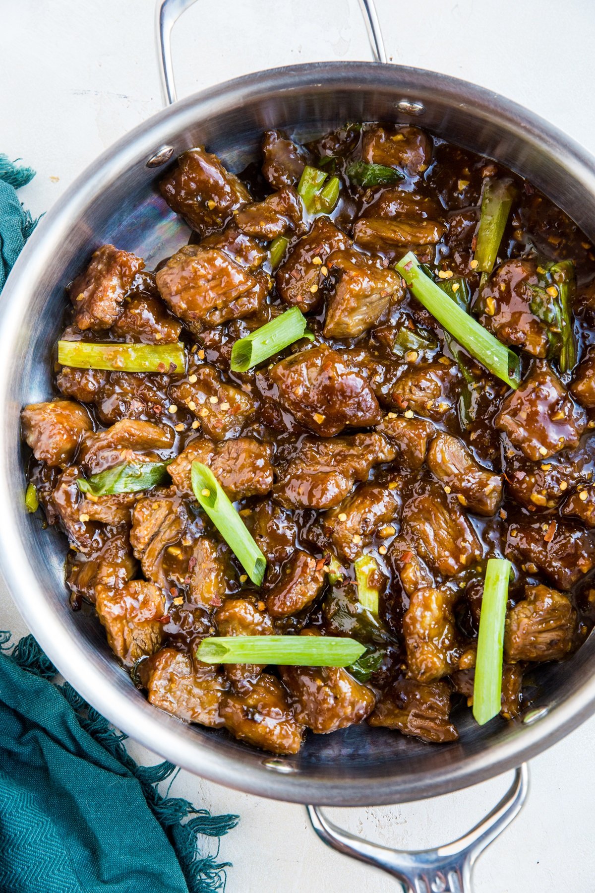 Stainless steel skillet full of healthy Mongolian beef with green onions on top and a blue napkin to the side.
