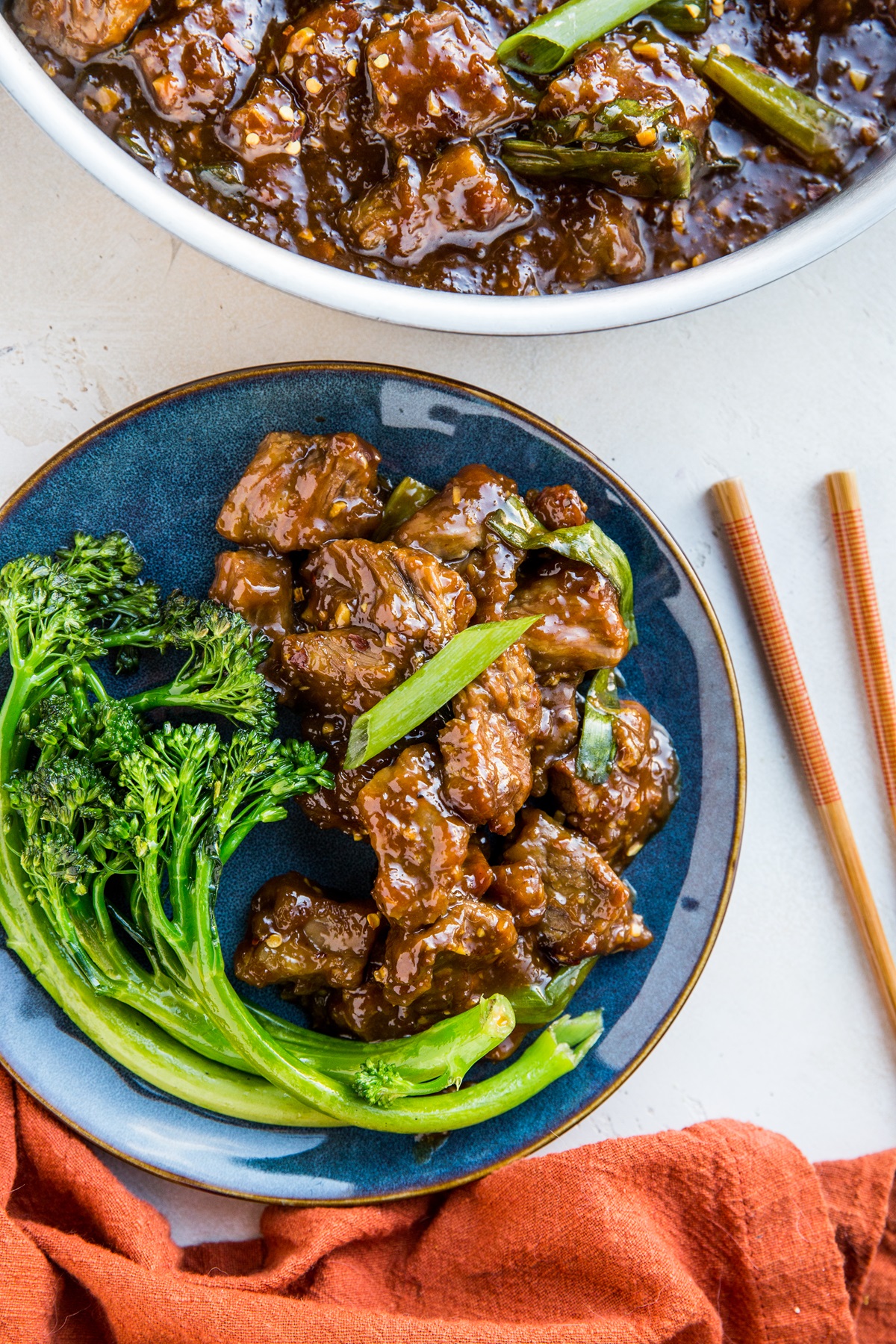 Blue plate of Mongolian beef with steamed broccolini and the skillet of the rest of the beef and a red napkin.