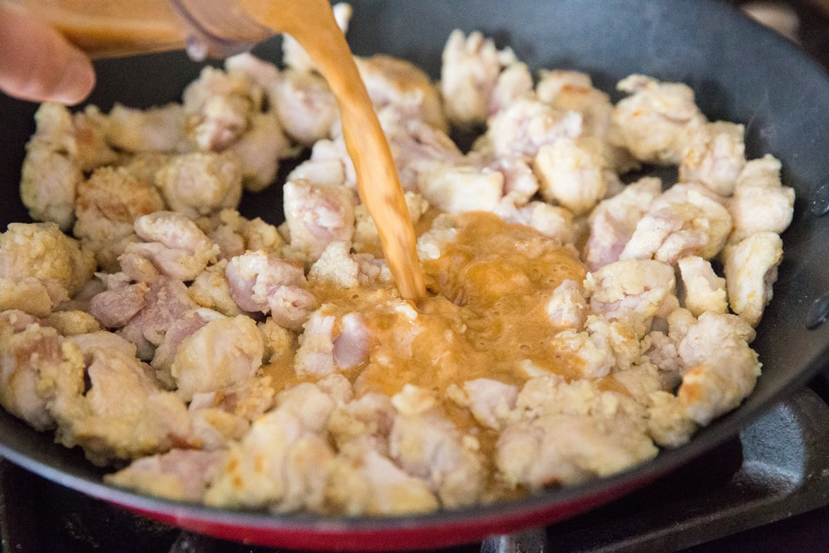 Pouring orange sauce into a skillet with browned chicken pieces.