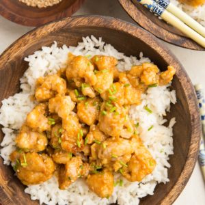 large wooden bowl full of rice and orange chicken with chop sticks and a red napkin.