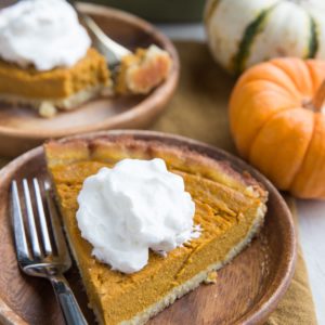 Slice of a Healthy Pumpkin Pie on a wooden plate with whipped cream on top with a wood inlay fork, ready to eat. Another slice in the background with fresh pumpkins to the side.