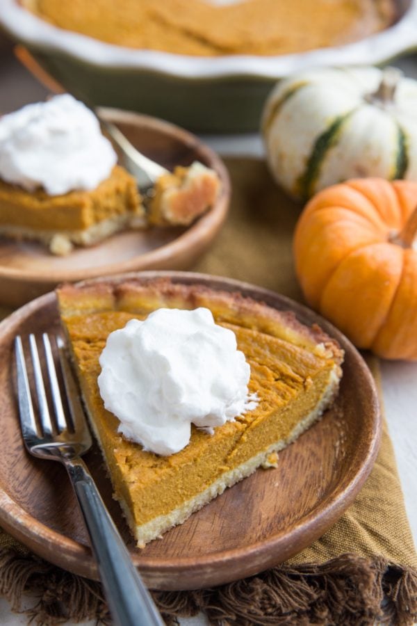 Slice of a Healthy Pumpkin Pie on a wooden plate with whipped cream on top with a wood inlay fork, ready to eat. Another slice in the background with fresh pumpkins to the side.