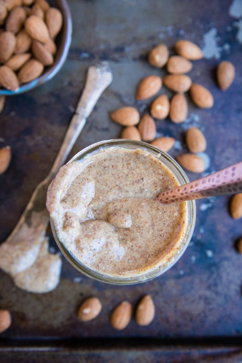 jar of homemade almond butter with a spoon inside of the jar and fresh almonds all around.