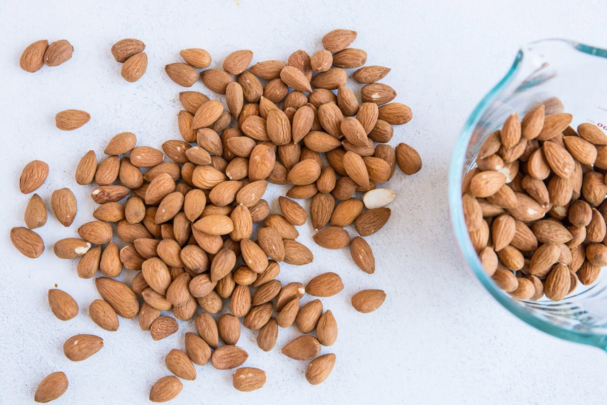 Raw almonds on a backdrop with a measuring cup full of almonds to the side.