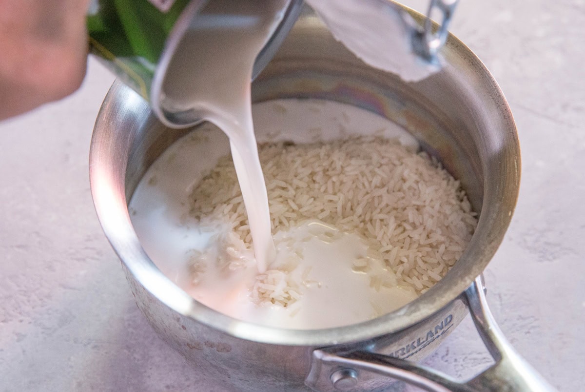 Hand pouring coconut milk into saucepan with rice to make coconut rice