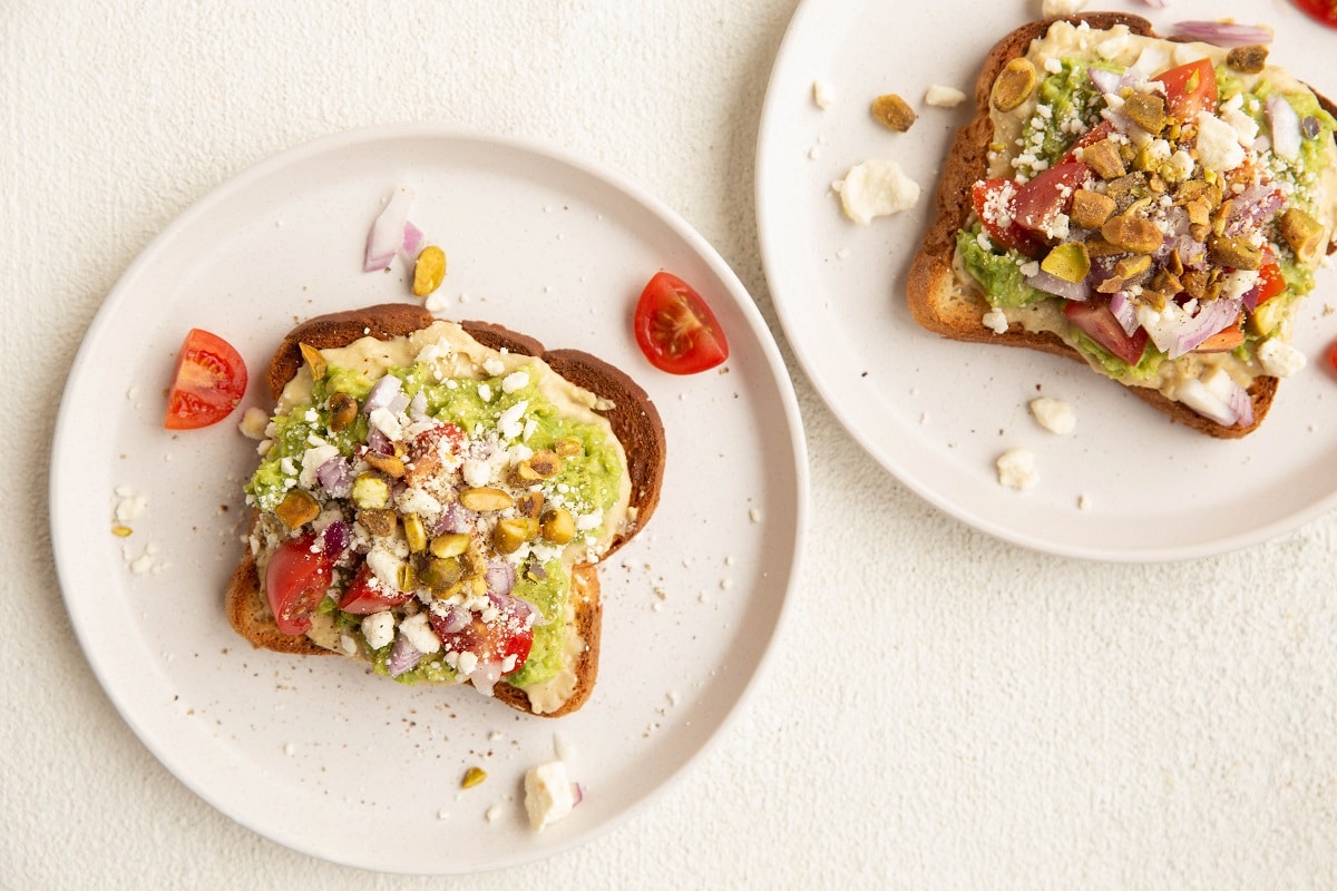 Two slices of hummus avocado toast with favorite toppings on two white plates.