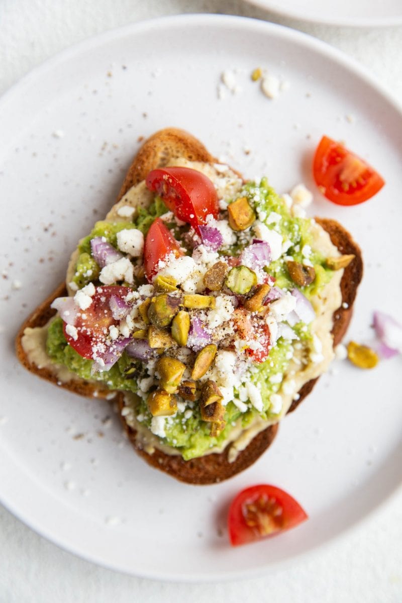 Close up top down image of avocado toast with hummus, tomatoes, red onion, feta, and pistachios.