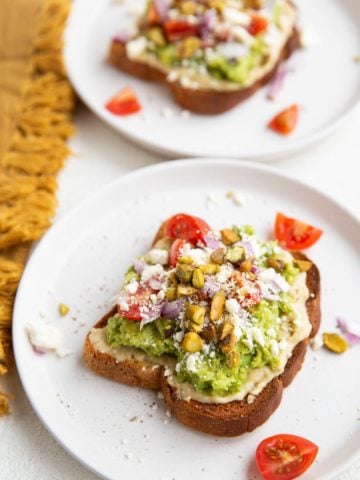 Two slices of avocado hummus toast on white plates, ready to eat.