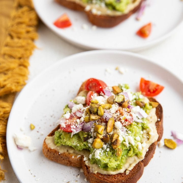 Two slices of avocado hummus toast on white plates, ready to eat.