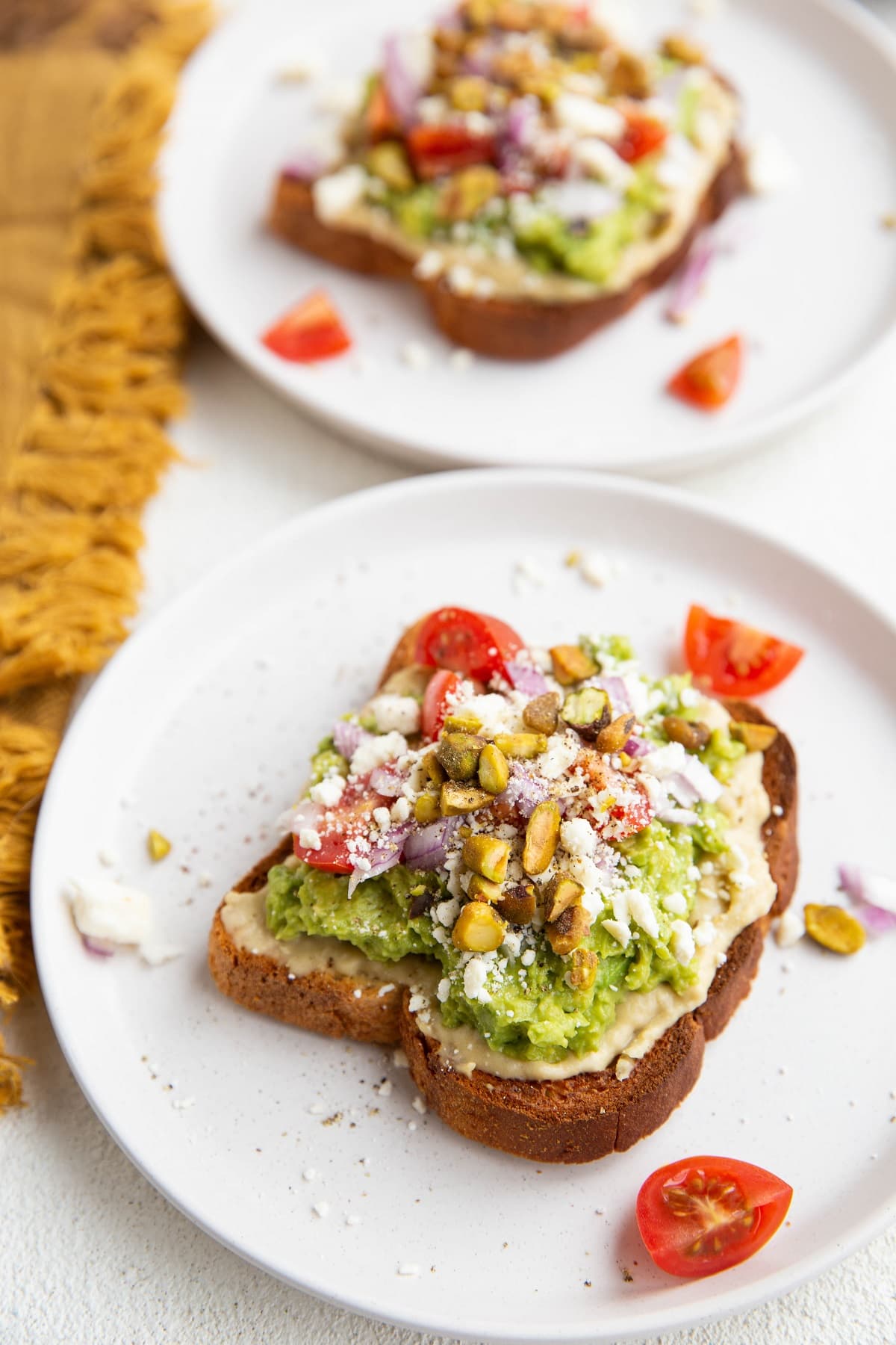 Two slices of avocado hummus toast on white plates, ready to eat.