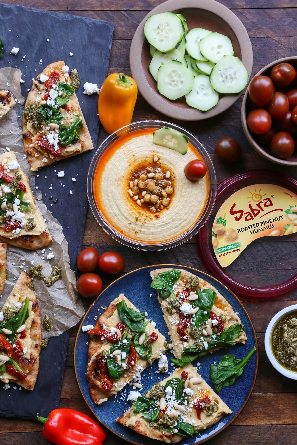 Hummus Flatbread with Sun-Dried Tomatoes, Spinach, and Pesto on a black slate with slices on a blue plate and hummus for dipping.