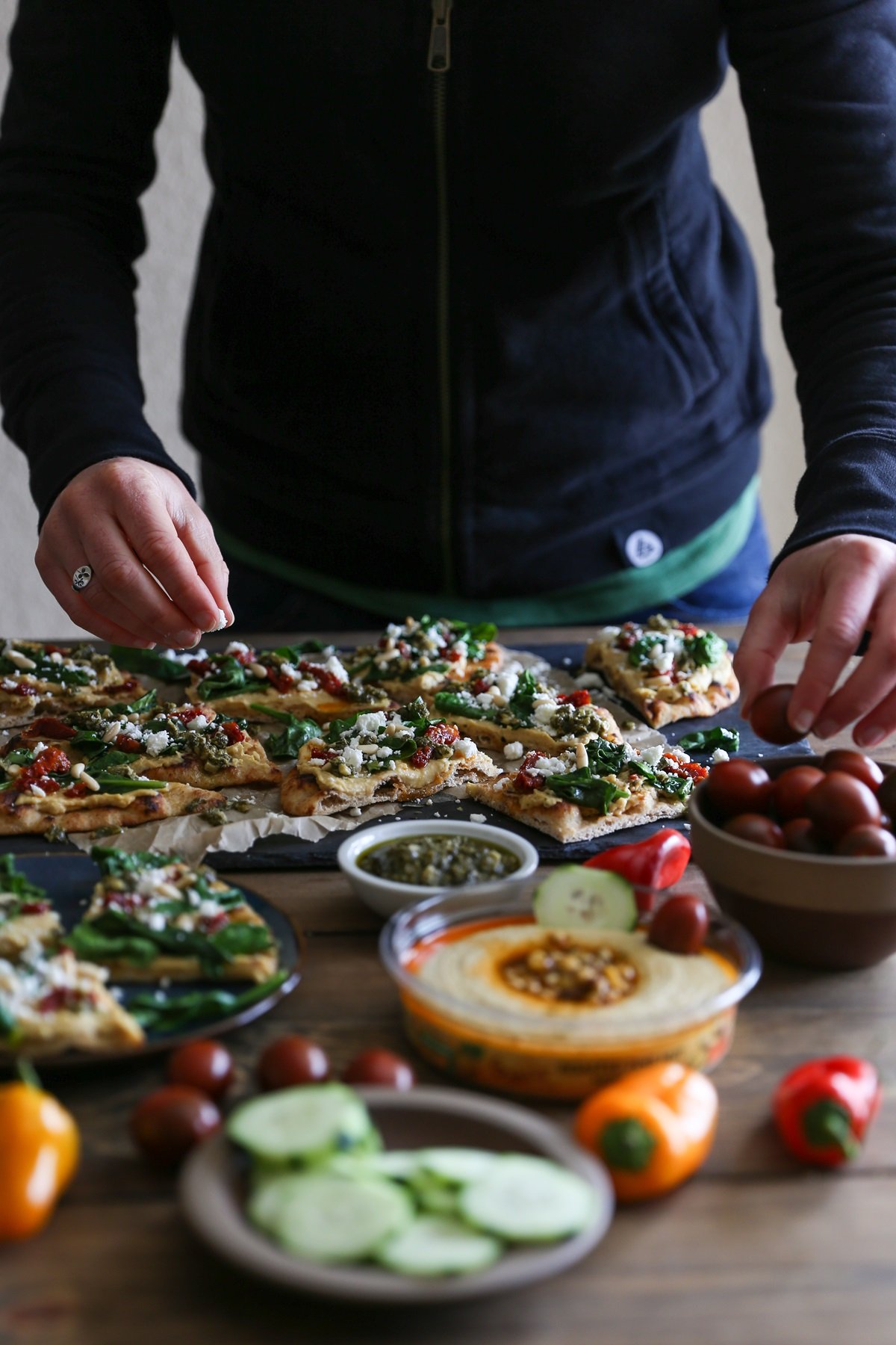 Woman topping hummus flatbread with toppings like tomatoes.