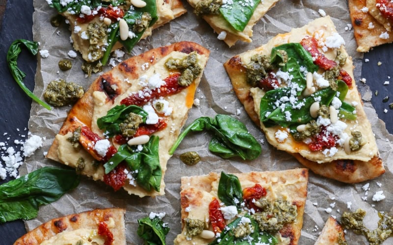 Black slate with slices of hummus flatbread on top, topped with sun-dried tomatoes, pesto sauce, spinach, and more.