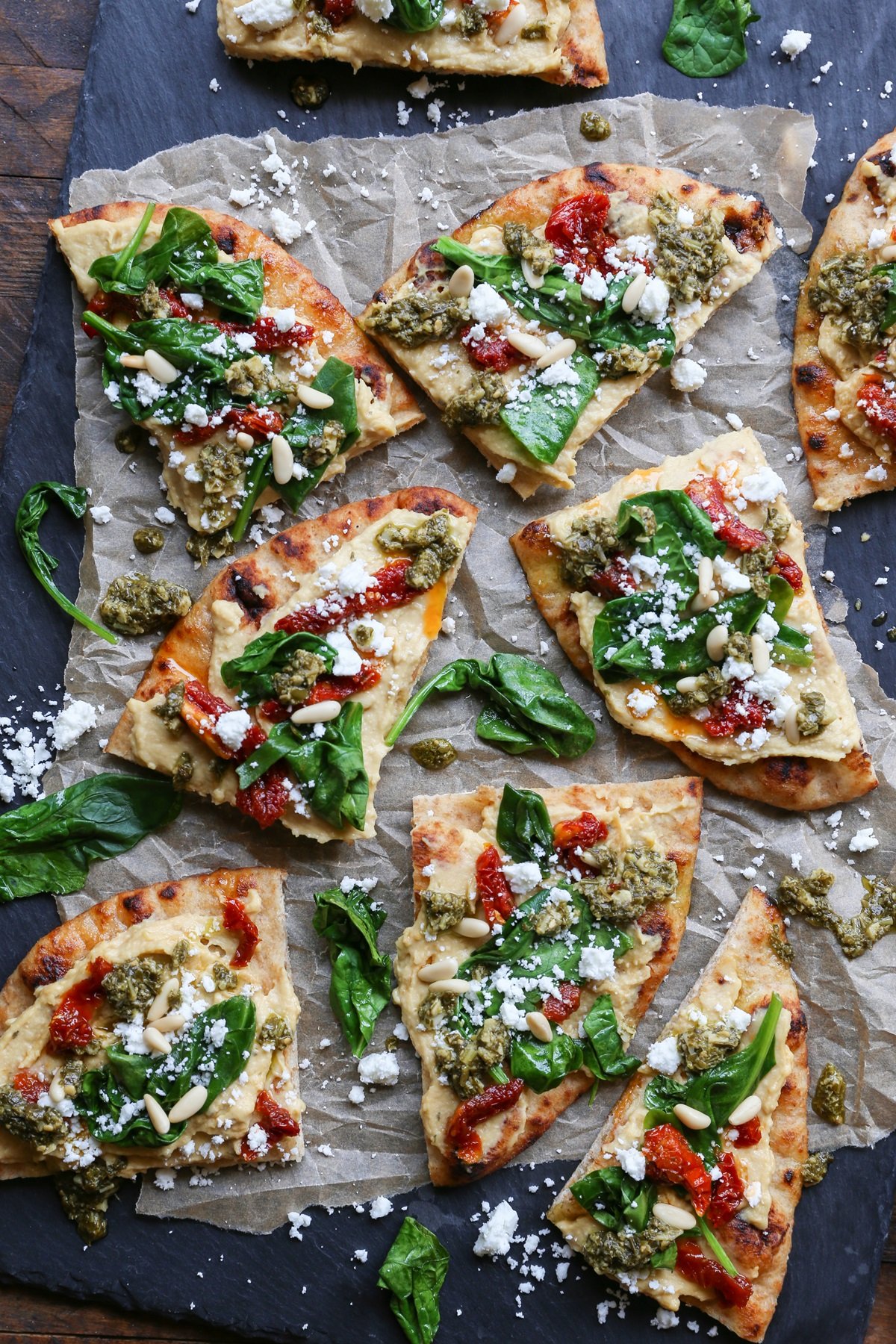 Black slate with slices of hummus flatbread on top, topped with sun-dried tomatoes, pesto sauce, spinach, and more.