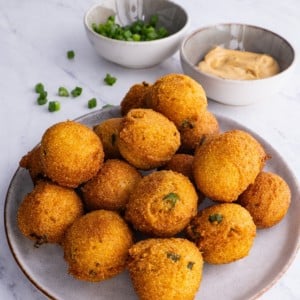 Plate full of hush puppies sprinkled with green onions with dipping sauce in the background.