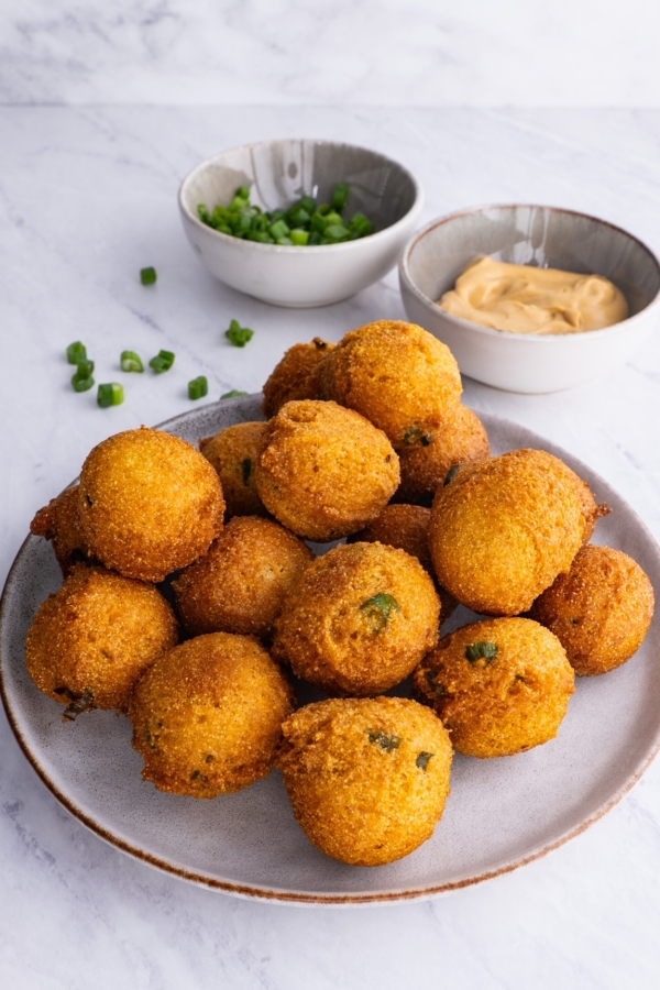 Plate full of hush puppies sprinkled with green onions with dipping sauce in the background.