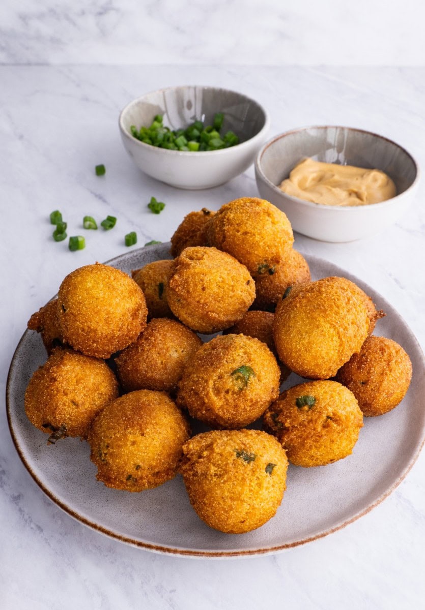 Plate full of hush puppies sprinkled with green onions with dipping sauce in the background.
