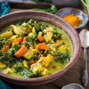 close up turmeric chicken soup in a brown bowl with a blue napkin to the side.