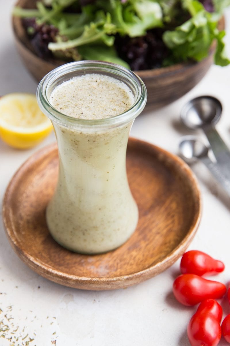Italian salad dressing in a glass container with fresh tomatoes to the side and a salad in the background.