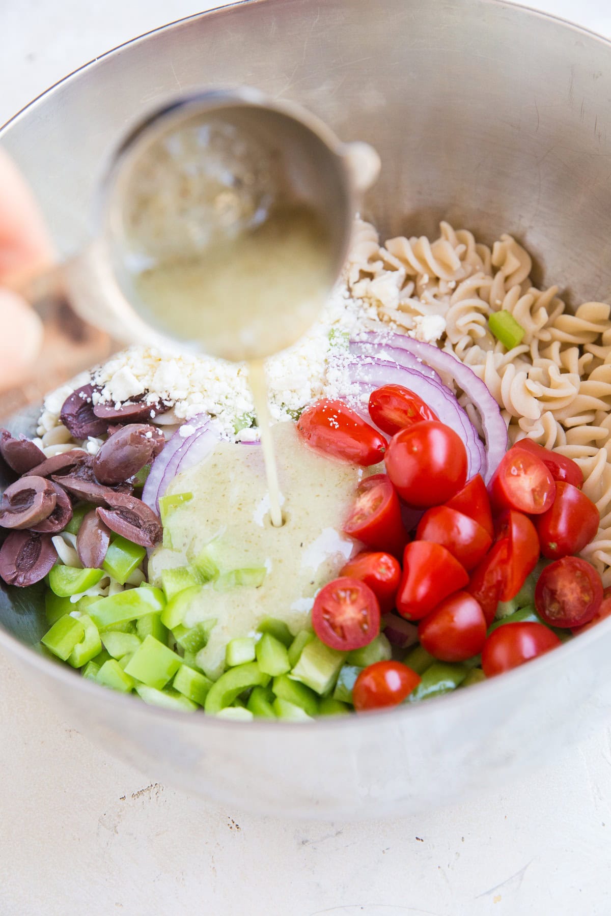 Large mixing bowl full of pasta salad ingredients with Italian dressing being poured in.
