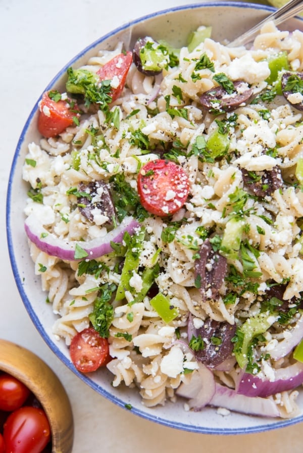 Gluten free Italian pasta salad in a large bowl with a bowl of cherry tomatoes to the side. Pasta noodles, red onions, bell peppers, cherry tomatoes, feta cheese, and more.