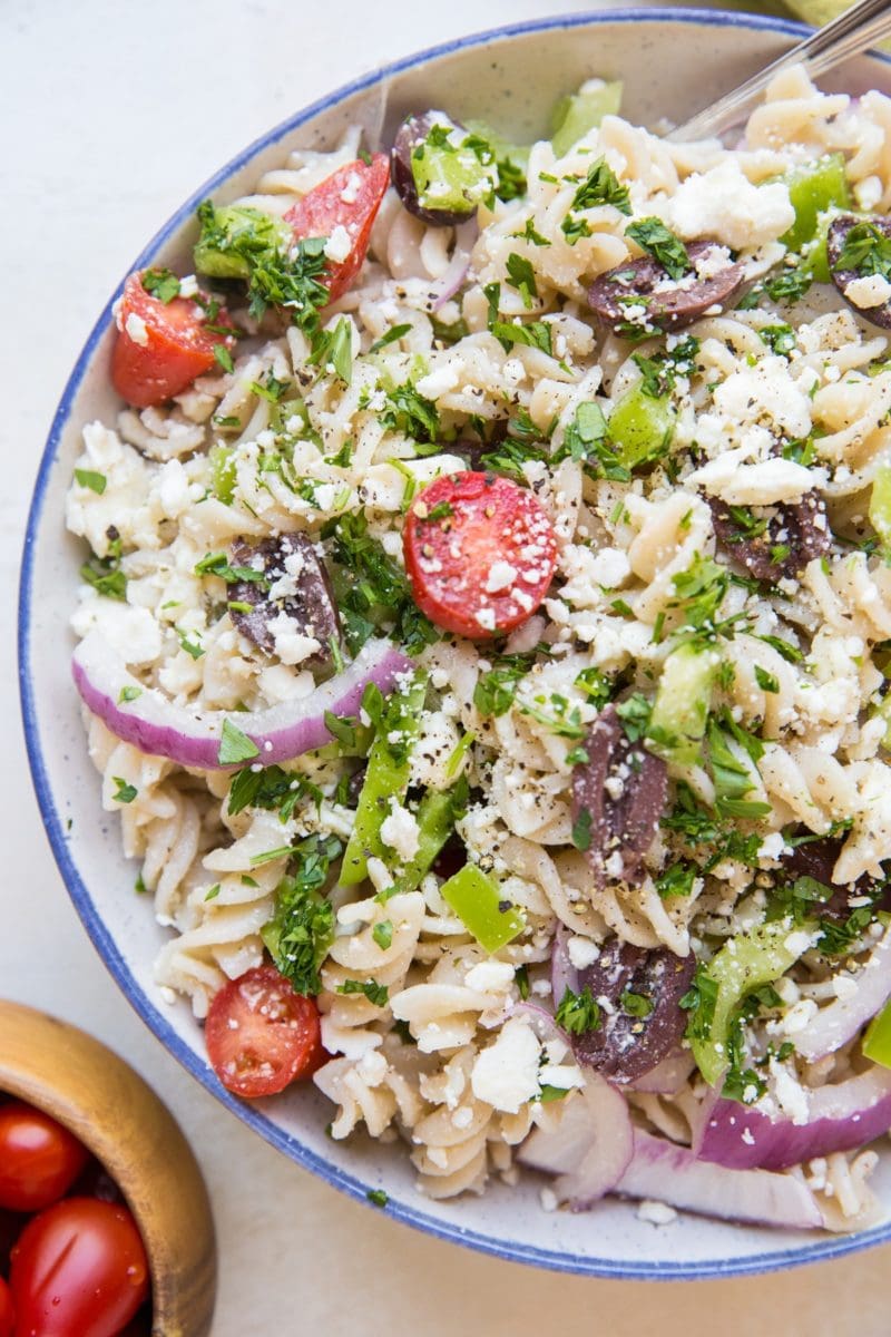 Gluten free Italian pasta salad in a large bowl with a bowl of cherry tomatoes to the side. Pasta noodles, red onions, bell peppers, cherry tomatoes, feta cheese, and more.