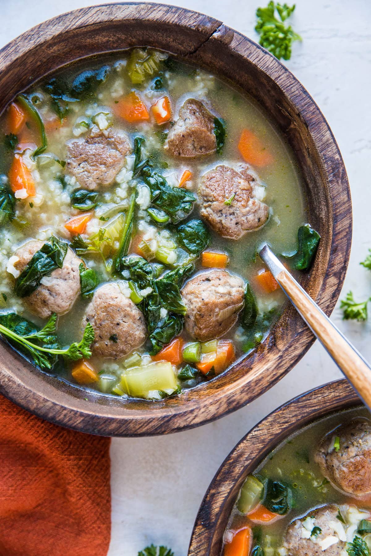 A wooden bowl filled with Italian wedding soup with rice, featuring meatballs, spinach, diced carrots, and celery. A spoon rests in the bowl while a second bowl and a rust-colored napkin are set nearby.