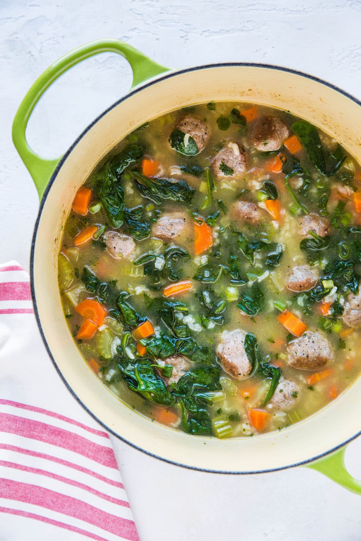 A green pot filled with Italian wedding soup with rice—meatballs, spinach, diced carrots, celery, and broth—set on a white surface next to a white cloth with pink stripes.