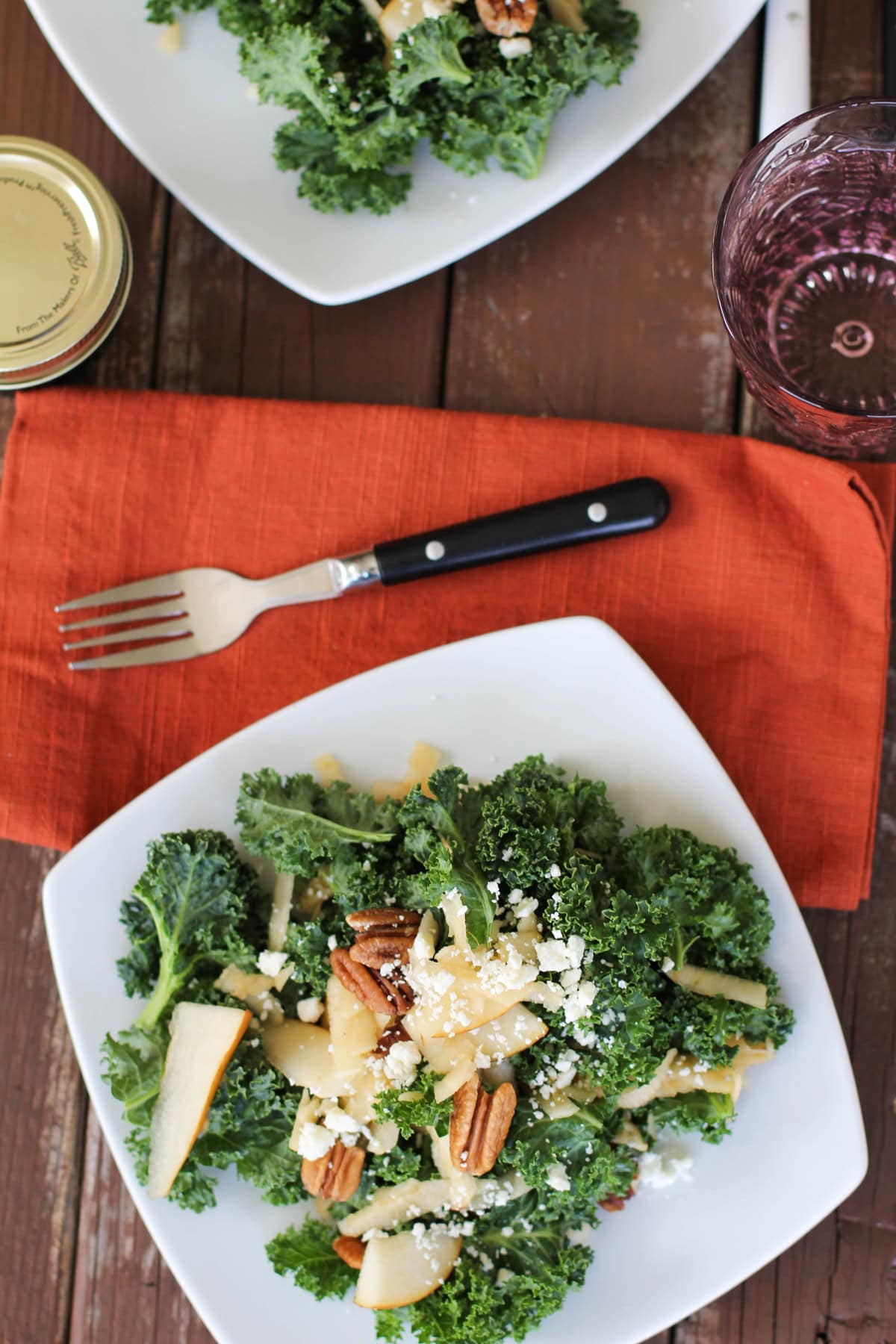 White plate of kale salad with pecans and blue cheese with a red napkin and a water glass to the side. Ready to serve.