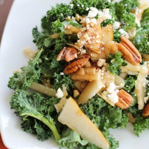 Massaged Kale Salad with Apple, Pear, Gorgonzola and Roasted Pecans on a white plate with a red napkin to the side.