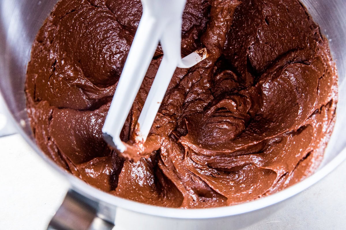 Thick, glossy keto brownies batter being mixed in a metal bowl with a white stand mixer paddle attachment.