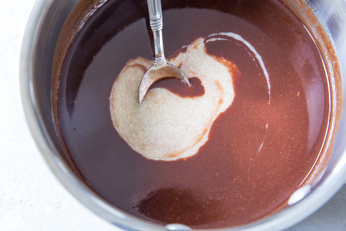 A metal spoon stirs a blend of light-colored peanut butter into smooth, glossy melted chocolate in a stainless steel bowl, the perfect base for rich keto brownies.