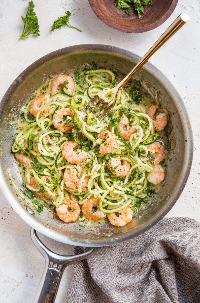 Stainless steel skillet full of zucchini noodles and shrimp with a fork twirling some of the noodles and a grey napkin to the side.