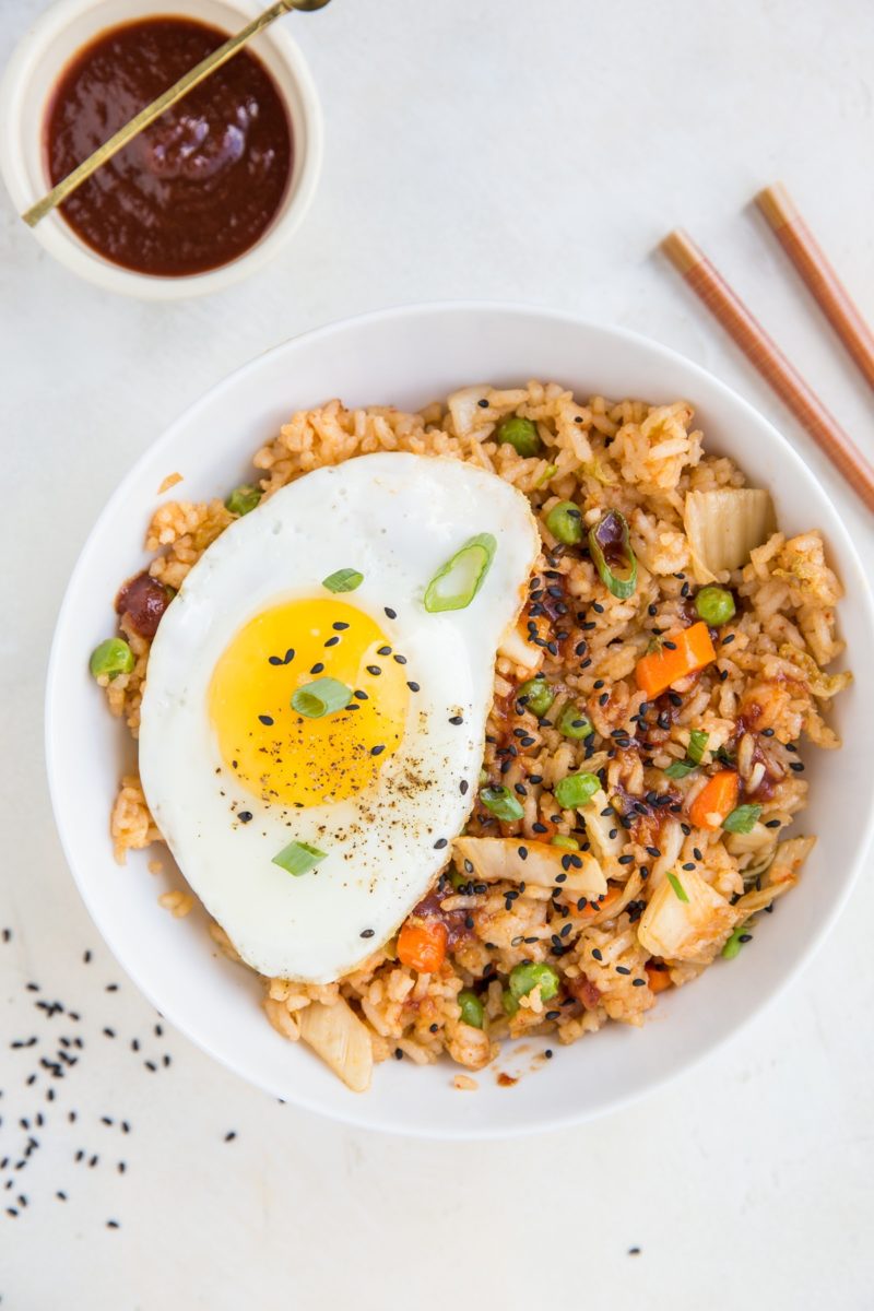 Large white bowl full of kimchi fried rice with a fried egg on top and sprinkles of sesame seeds to the side.