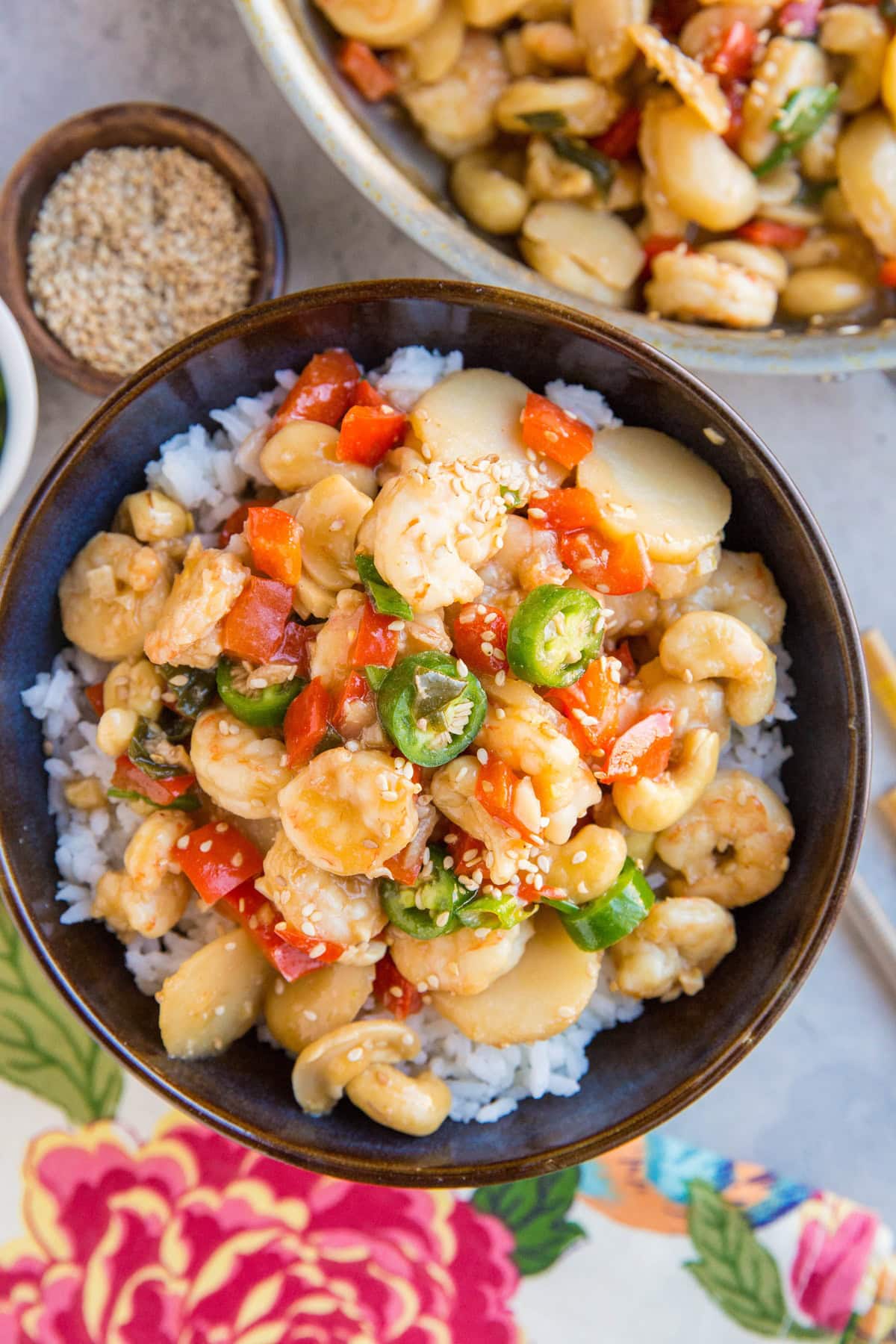 Kung pao shrimp on top of rice in a blue ceramic bowl with a floral napkin to the side and the skillet with the rest of the shrimp to the side.