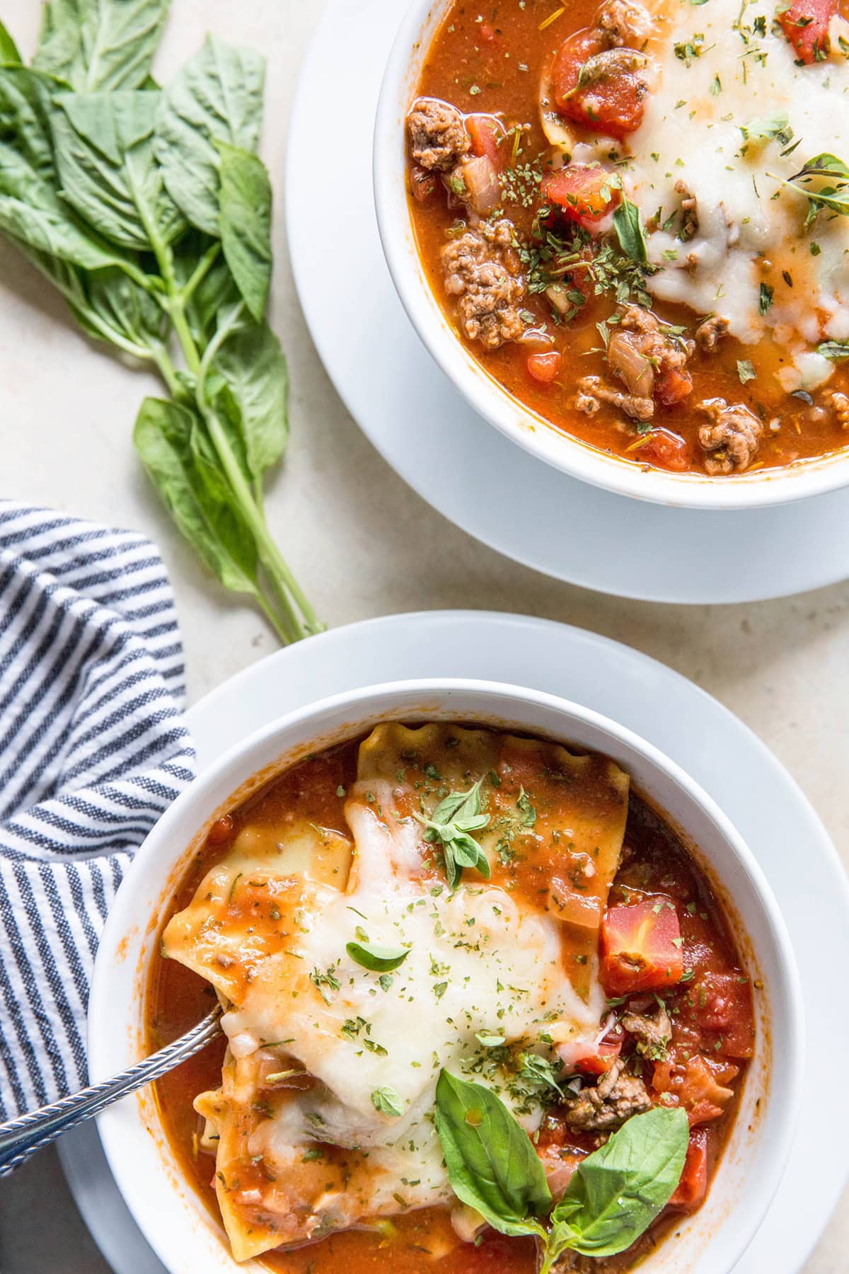 Two bowls of lasagna soup with a spoon inside of one of them, ready to serve. Fresh basil leaves and dried parsley sprinkled on top.