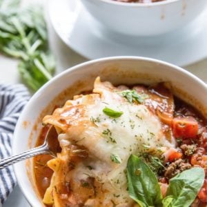 Lasagna soup in a white bowl with a spoon scooping some of the noodles and cheese out. A fresh basil leaf to the side and another bowl of soup in the background.
