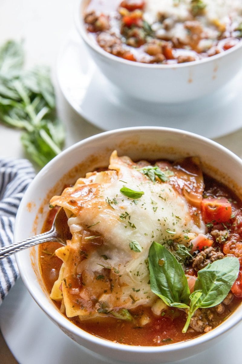 Lasagna soup in a white bowl with a spoon scooping some of the noodles and cheese out. A fresh basil leaf to the side and another bowl of soup in the background.