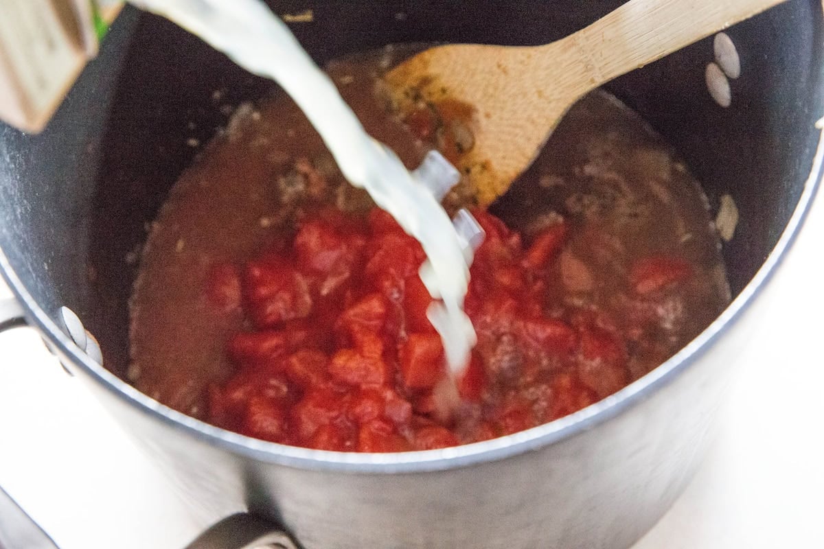 Pouring chicken broth into a pot with diced tomatoes and beef.