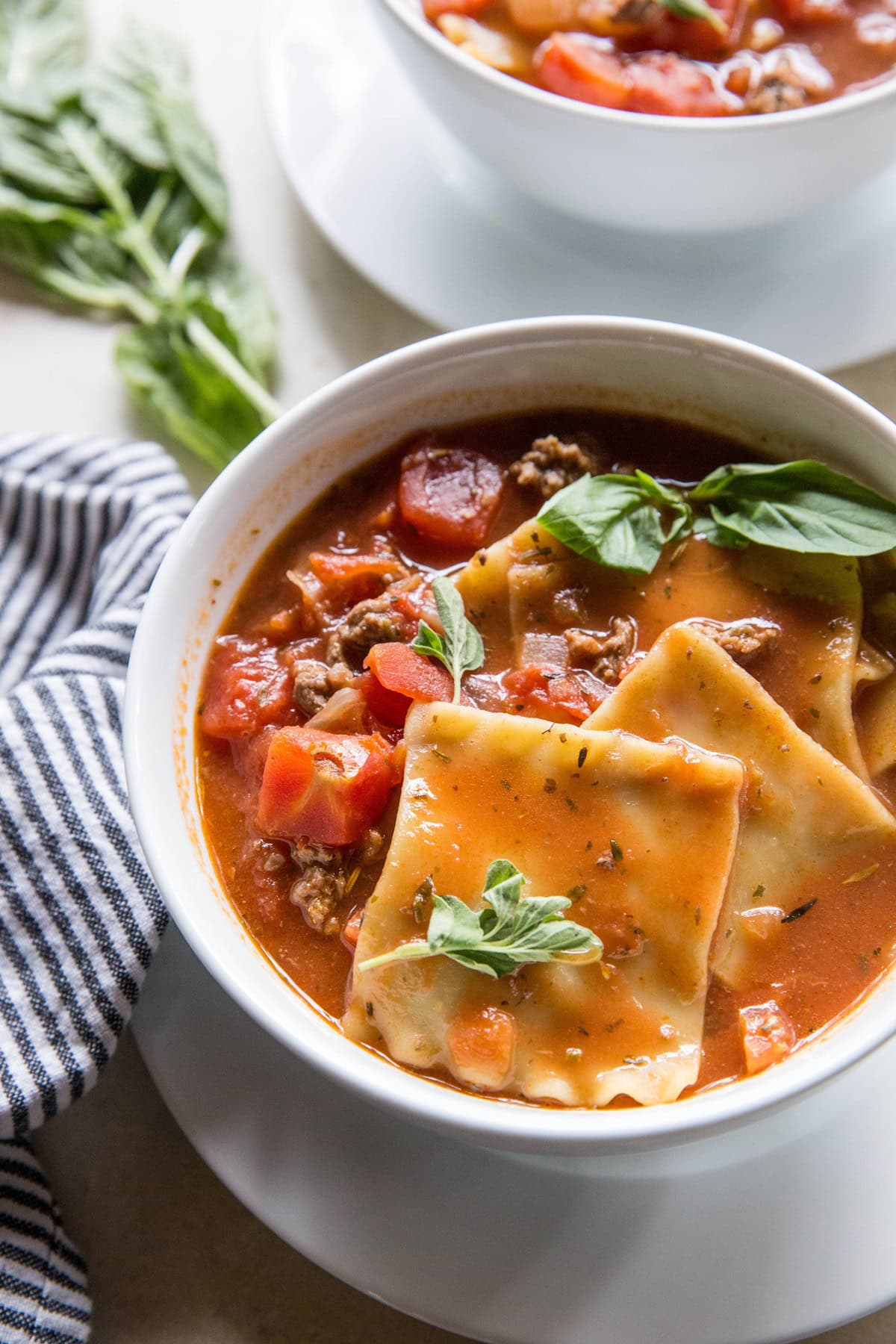 Two white bowls of lasagna soup, garnished with basil with a blue striped napkin