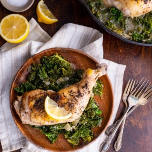 Wooden plate with Lemony Braised Chicken with Kale and the cast iron skillet with the rest of the chicken and kale. Napkins and forks around for serving.