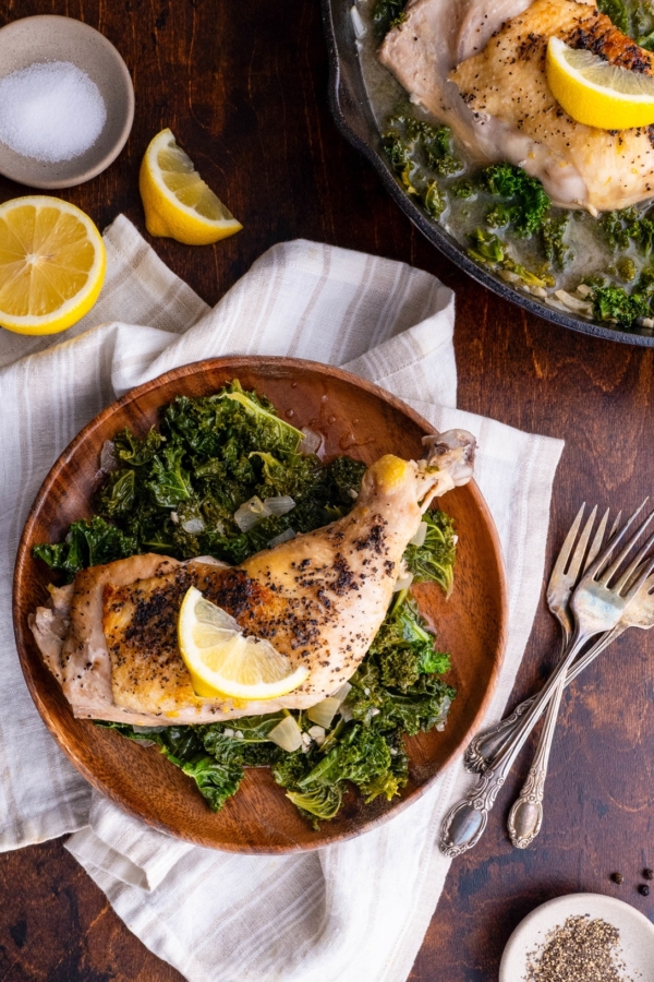 Wooden plate with Lemony Braised Chicken with Kale and the cast iron skillet with the rest of the chicken and kale. Napkins and forks around for serving.