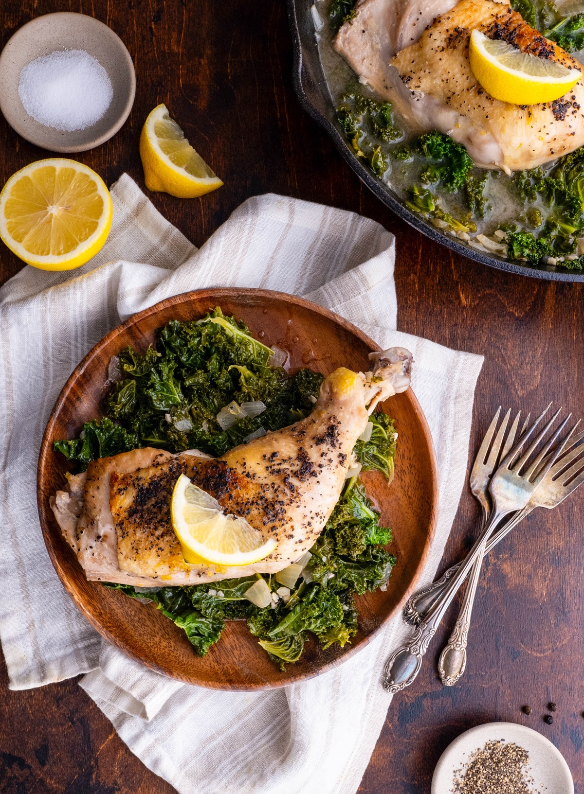 Wooden plate with Lemony Braised Chicken with Kale and the cast iron skillet with the rest of the chicken and kale. Napkins and forks around for serving.