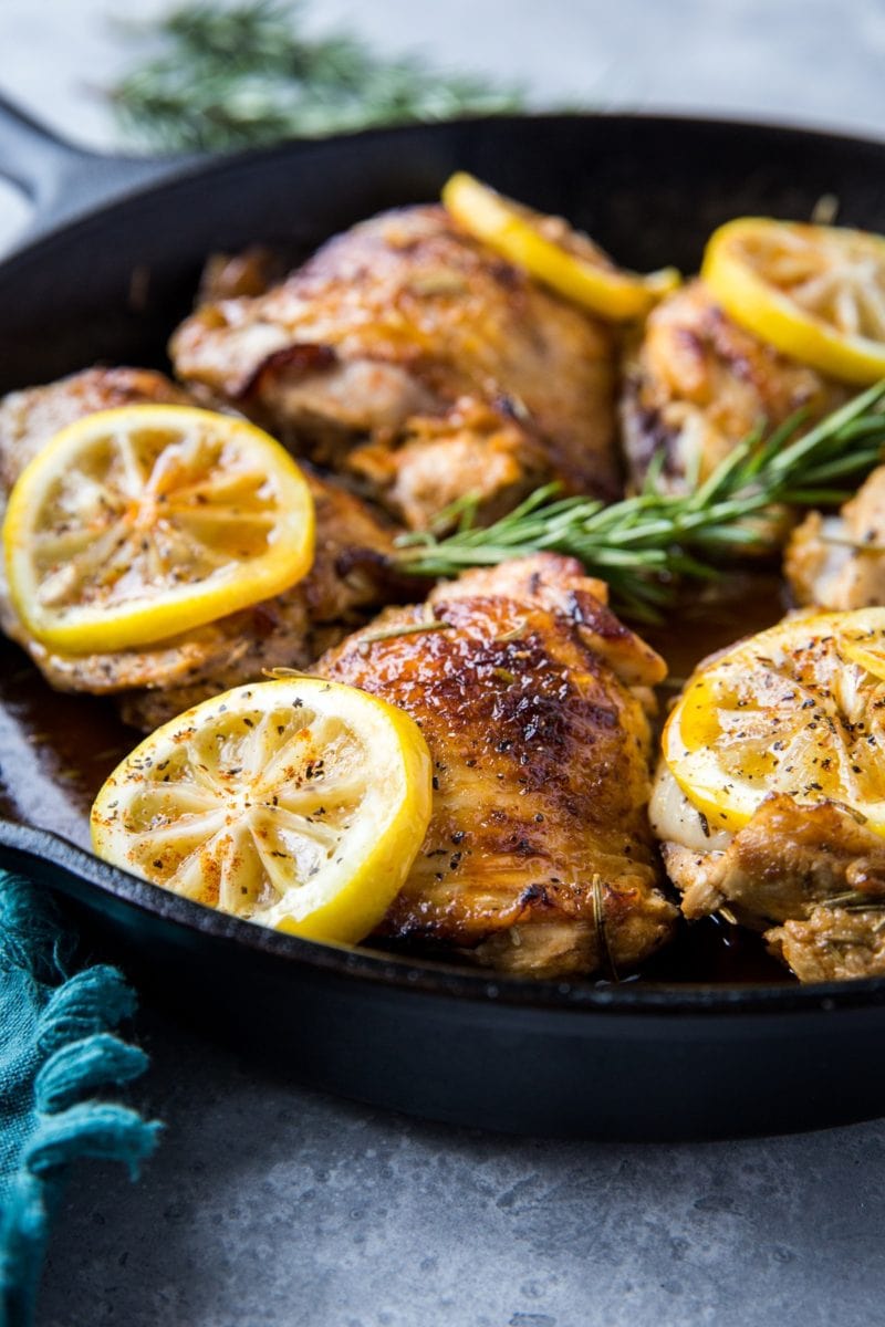 Braised skin-on chicken thighs in a cast iron skillet with fresh rosemary and lemon slices.