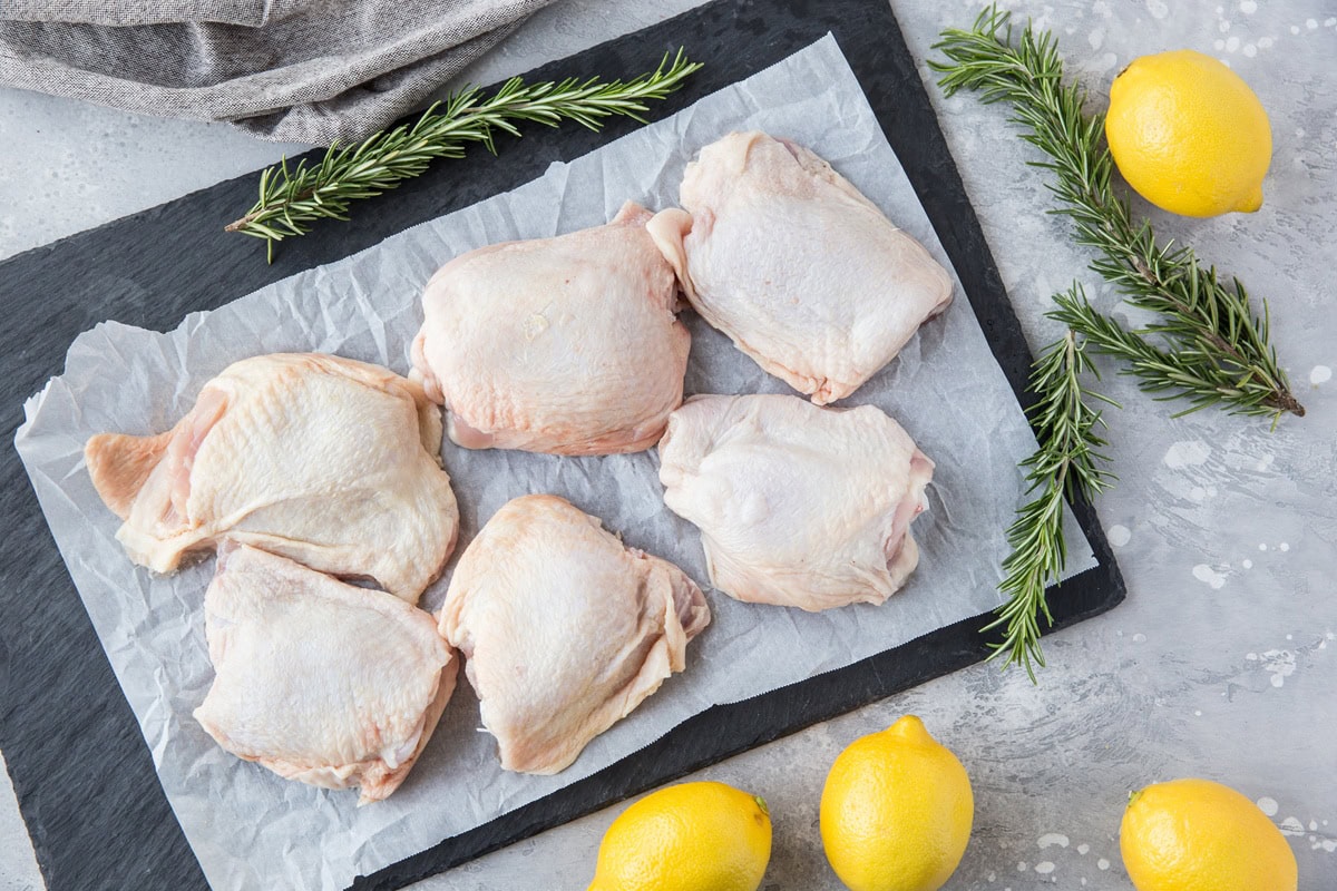 Black slate cutting board with raw chicken thighs on top and fresh lemons to the side with fresh rosemary. Ready to make a chicken thigh recipe.