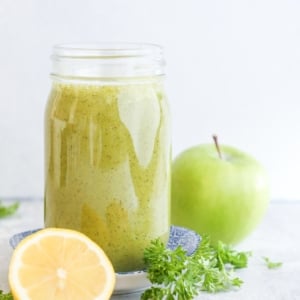 Large glass of smoothie with a green apple in the background, fresh parsley, a lemon and turmeric.