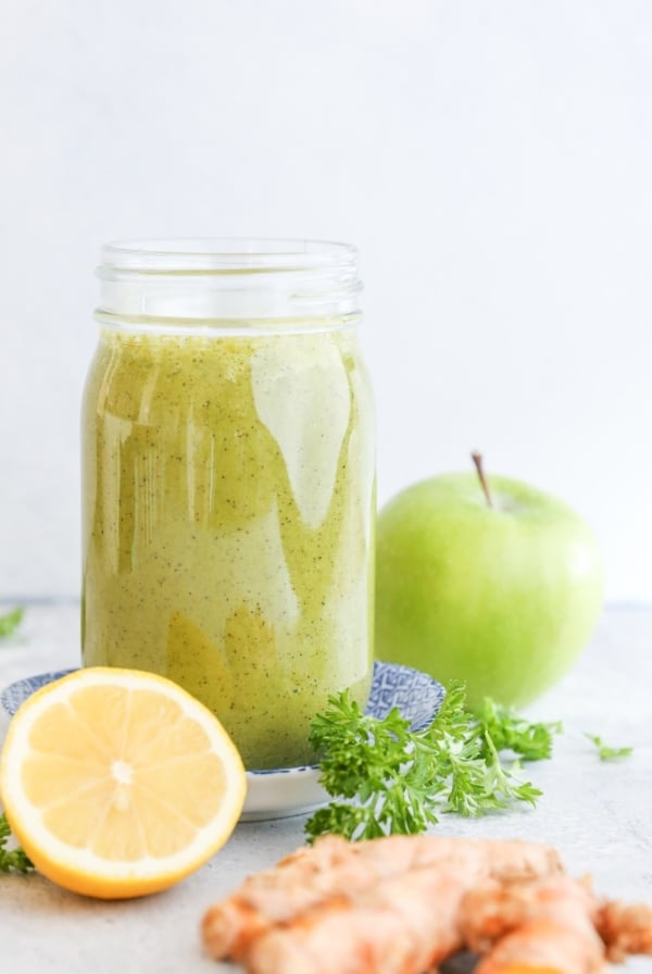 Large glass of smoothie with a green apple in the background, fresh parsley, a lemon and turmeric.