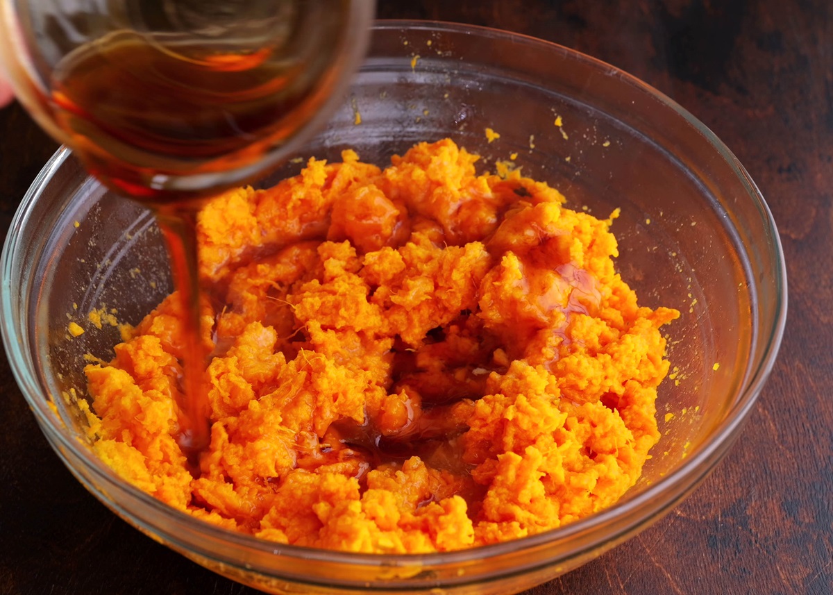Glass bowl full of mashed sweet potatoes with pure maple syrup being poured in.