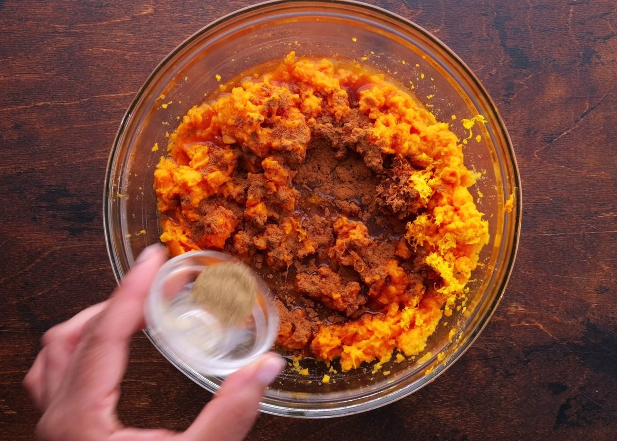 Glass bowl of mashed sweet potatoes with maple syrup, cinnamon, and cardamom being poured in.