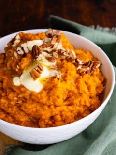 Mashed sweet potatoes in a white bowl with melted butter on top and chopped pecans. A teal tea towel to the side.